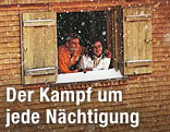 Zwei Personen schauen bei Schneefall aus dem Fenster eines alpinen Holzhauses