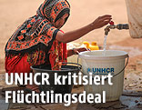 Ein weiblicher Flüchtling füllt Wasser in einen Kübel mit UNHCR-Zeichen